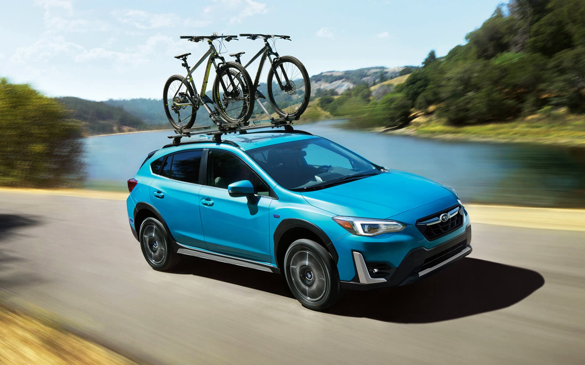 A blue Crosstrek Hybrid with two bicycles on its roof rack driving beside a river | John Kennedy Subaru in Plymouth Meeting PA